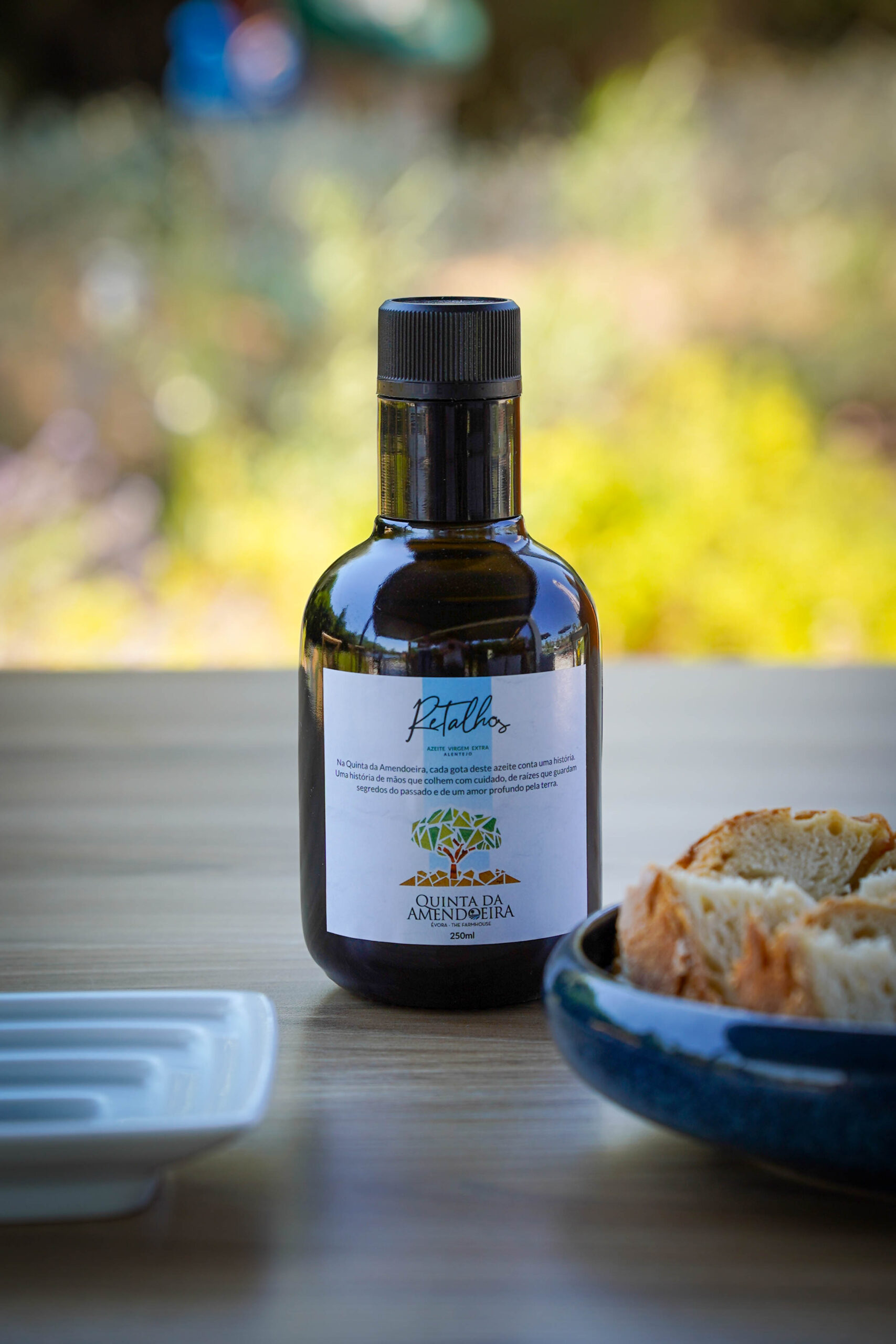 Dark glass olive oil bottle with a black ridged cap on a wooden table, label reads Quinta da Amendoeira, with bread nearby in the foreground or right edge.