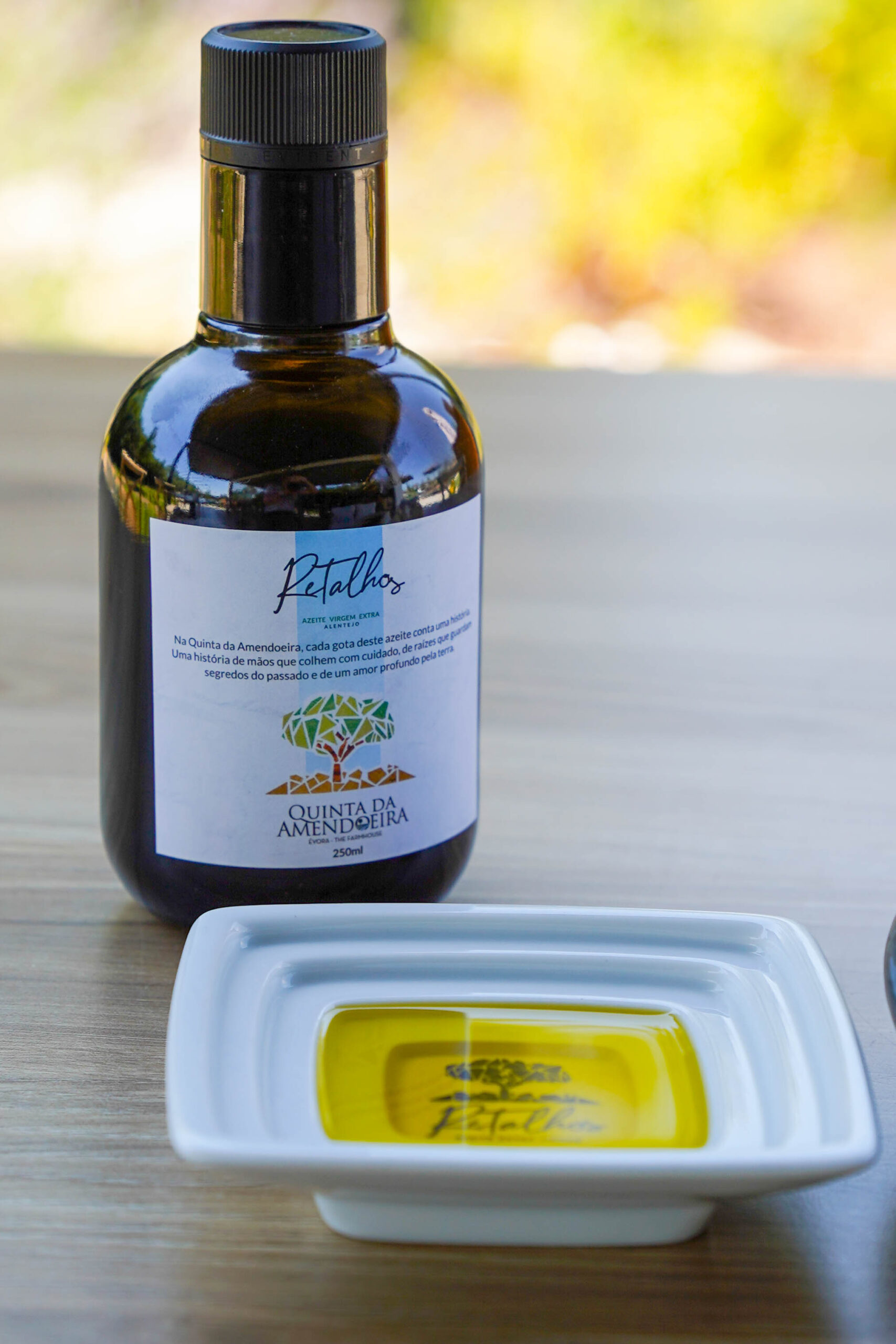 Close-up of a dark glass olive oil bottle with a black cap on a wooden table; label shows tree artwork and Portuguese text Quinta da Amendoeira.