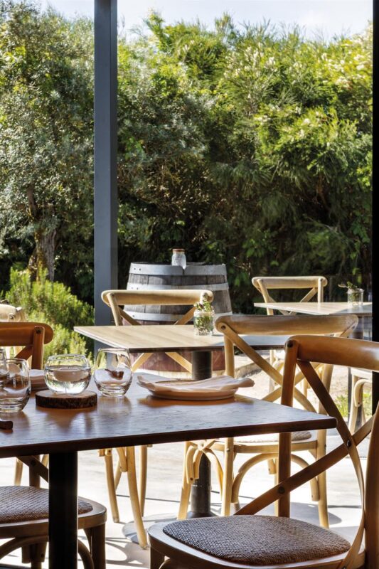 Outdoor cafe seating with wooden tables and chairs, glassware, and lush green garden beyond the patio paneling.
