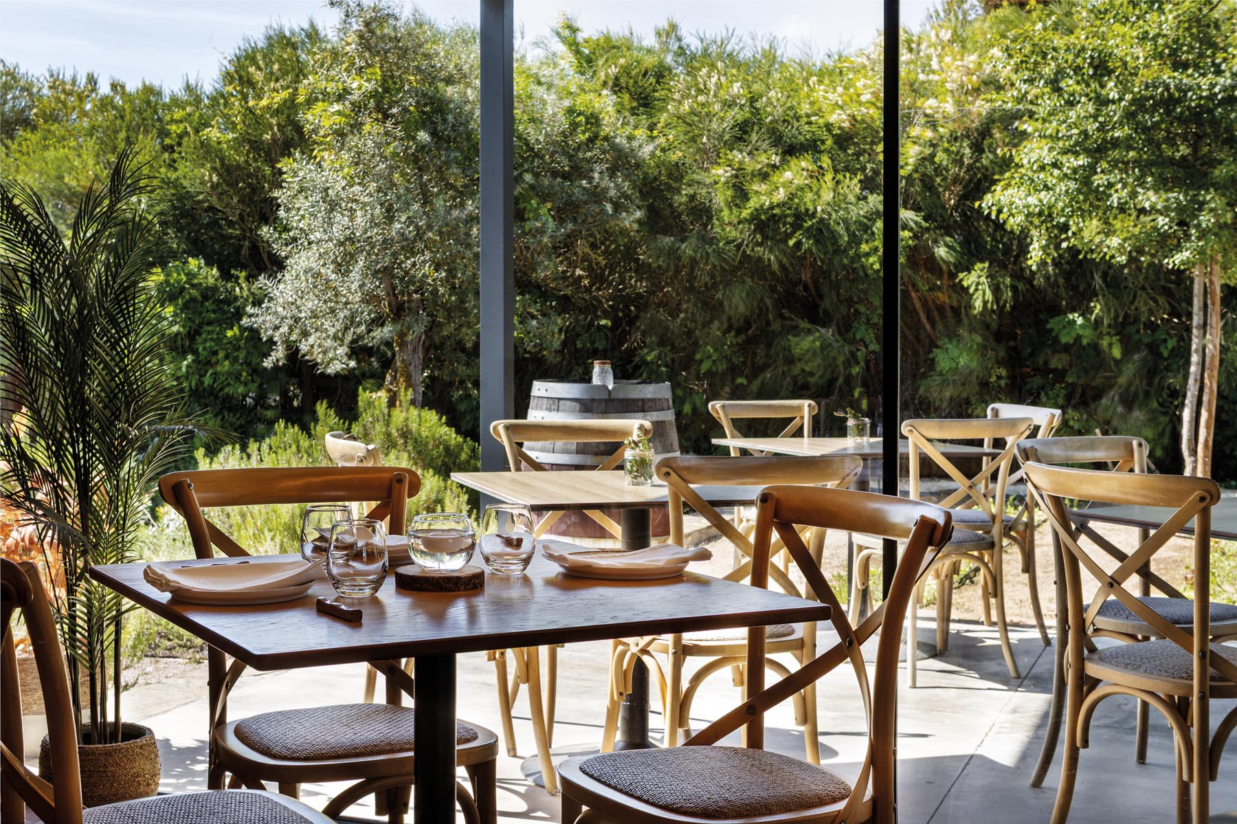 Outdoor cafe seating with wooden tables and chairs, glassware, and lush green garden beyond the patio paneling.