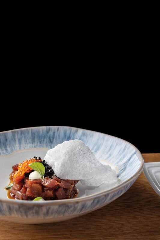 Tuna tartare in a blue-rimmed bowl with a crisp white shard, on a wooden table, against a black background; sauce bottle in the background to the right.