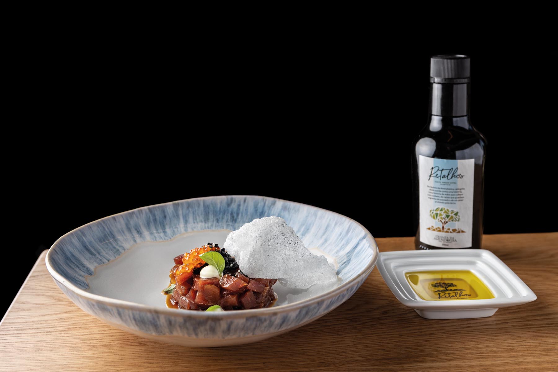 Tuna tartare in a blue-rimmed bowl with a crisp white shard, on a wooden table, against a black background; sauce bottle in the background to the right.