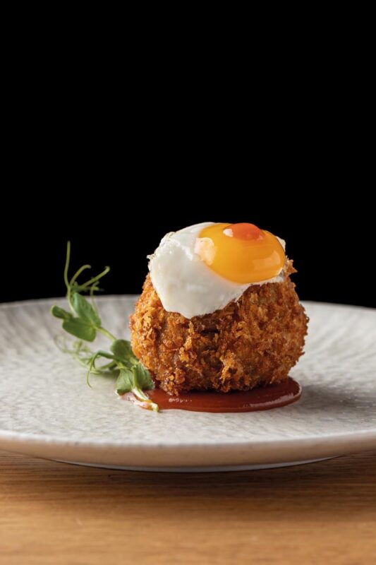 Crispy croquette topped with a sunny-side-up egg on a drizzle of red sauce, garnished with microgreens on a gray plate.
