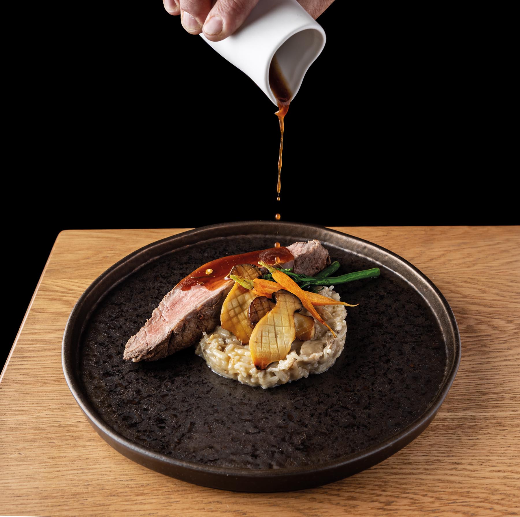 Pouring brown sauce onto a plated beef dish with vegetables and risotto on a dark plate.