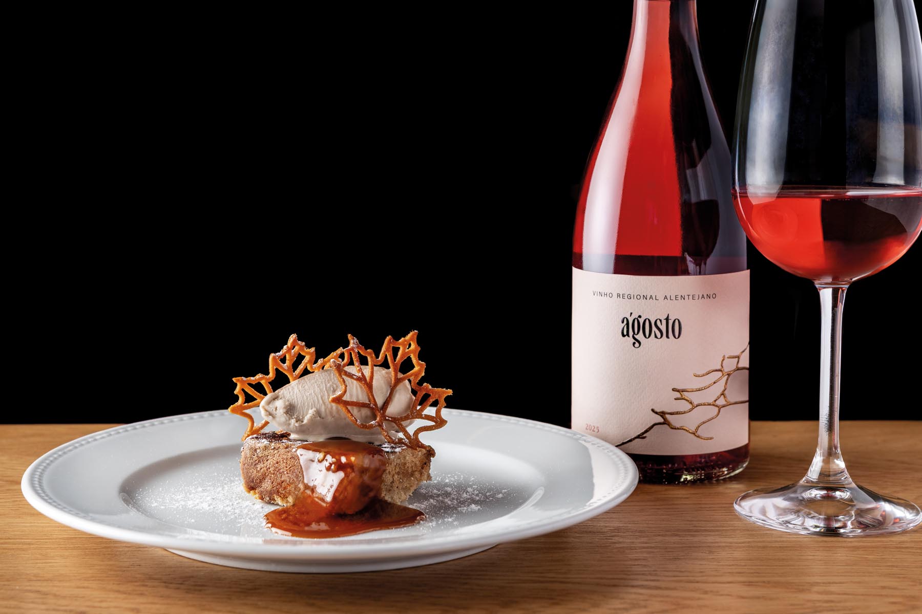 Dessert on a white plate with caramel lattice tuile and red sauce beside a bottle of wine and a red wine glass on a wooden table against a black background.